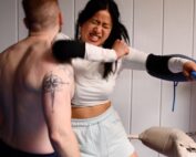 In einem Boxring mit Seilen und weißen Polstern schlägt eine Frau in weißem Hemd und hellen Shorts auf einen tätowierten Mann ein, der kein Hemd trägt. Ihr langes schwarzes Haar weht, während sie Schutzkleidung an ihren Handgelenken trägt. Der männliche Gegner ist abgewandt und verkörpert die Stärke, die man bei Sportveranstaltungen der Hochschule Coburg oft sieht.