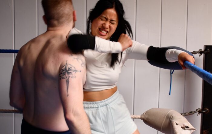 In einem Boxring mit Seilen und weißen Polstern schlägt eine Frau in weißem Hemd und hellen Shorts auf einen tätowierten Mann ein, der kein Hemd trägt. Ihr langes schwarzes Haar weht, während sie Schutzkleidung an ihren Handgelenken trägt. Der männliche Gegner ist abgewandt und verkörpert die Stärke, die man bei Sportveranstaltungen der Hochschule Coburg oft sieht.