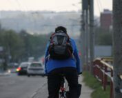 Eine Person mit einer blauen Jacke und einem schwarzen Rucksack fährt an einem bewölkten Tag mit dem Fahrrad die Straße in der Nähe der Hochschule Coburg entlang. Im Hintergrund sind Autos und Gebäude zu sehen.