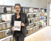 Ein Mann mit braunen Haaren und grauem Bart steht vor den Bücherregalen der Hochschule Coburg und hält ein Buch mit dem Titel „50 Jahre KHM: MAMIT“ in der Hand. Die Regale sind ordentlich mit Büchern gefüllt, in einem hellen Raum mit großem Fenster.