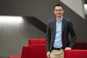 A person in a blue shirt and dark blazer stands confidently, smiling in a modern office space with red seating, embodying the professional spirit of Hochschule Coburg.