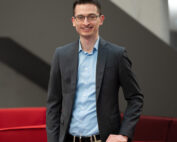 Eine Person mit Brille, grauem Blazer, hellblauem Hemd und Khakihose steht lächelnd neben einem roten Sofa an der Hochschule Coburg. Im Hintergrund ist eine graue Wand zu sehen.