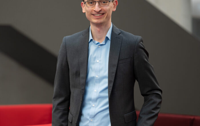 Eine Person mit Brille, grauem Blazer, hellblauem Hemd und Khakihose steht lächelnd neben einem roten Sofa an der Hochschule Coburg. Im Hintergrund ist eine graue Wand zu sehen.