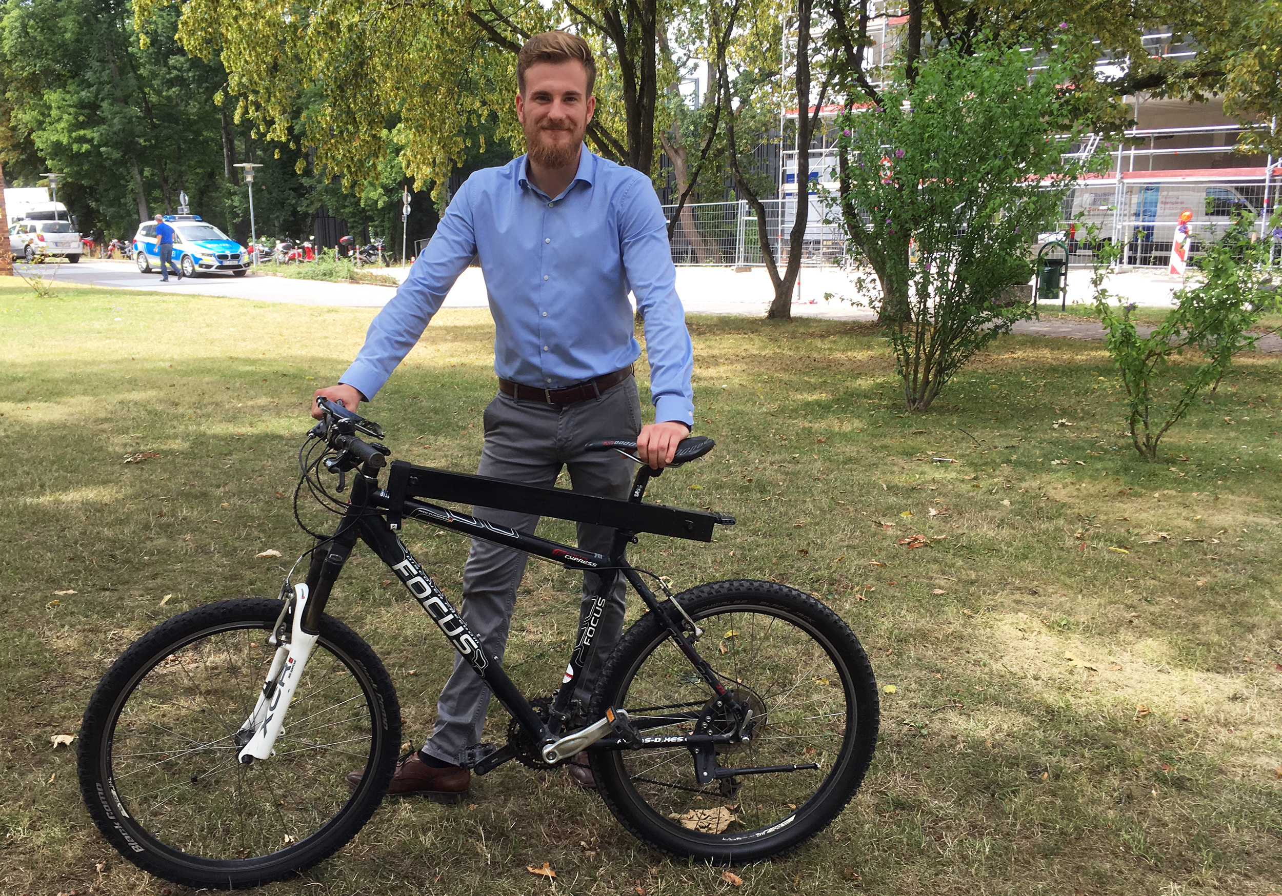 Ein bärtiger Mann in blauem Hemd und grauer Hose steht lächelnd in einer grünen Parkanlage in der Nähe der Hochschule Coburg und hält ein schwarzes Mountainbike in der Hand. Hinter ihm sind Bäume und ein Gebäude im Bau.