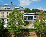 Eine malerische Ansicht des Campus der Hochschule Coburg zeigt links ein historisches Steingebäude und in der Mitte ein modernes weißes Gebäude. Eine Person schlendert unter einem teilweise bewölkten Himmel zwischen Bäumen und gepflegtem Gras den Weg entlang.
