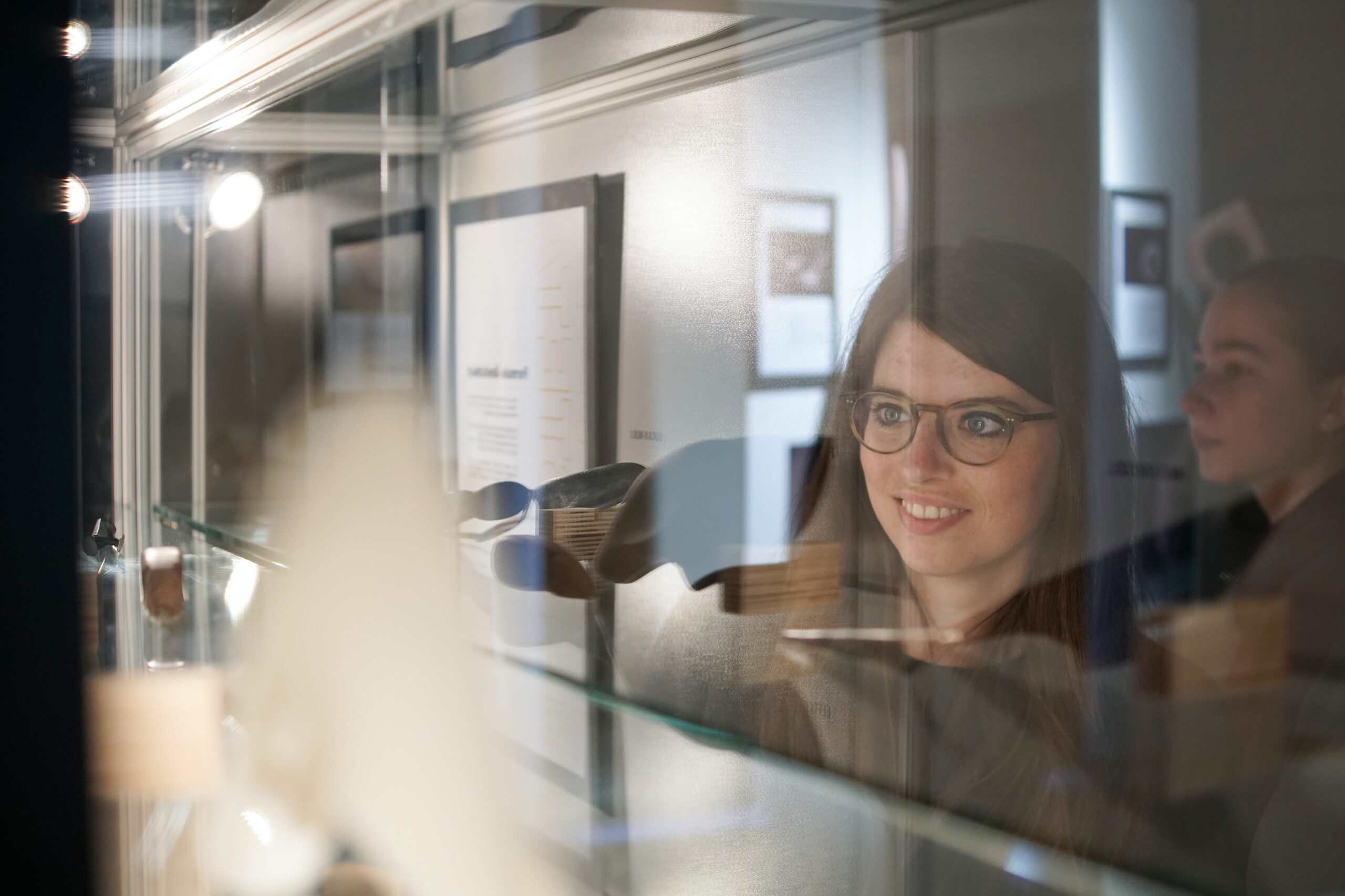 Eine Person mit Brille und langen Haaren lächelt, während sie Objekte in einer Glasvitrine in der Ausstellung der Hochschule Coburg bewundert. Ihr Spiegelbild fängt ihren entzückten Gesichtsausdruck ein, während ein anderer Besucher im Hintergrund verschwommen zu sehen ist. Die Umgebung strahlt eine Atmosphäre stiller Neugier und Gelehrsamkeit aus.