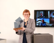 Eine Person steht in einem Innenbereich der Hochschule Coburg und hält lächelnd eine transparente Auszeichnung in den Händen. Im Hintergrund ist auf einem Bildschirm eine Videokonferenz mit vier Teilnehmern zu sehen. Auf einem Podest in der Nähe ist eine weitere Auszeichnung ausgestellt.