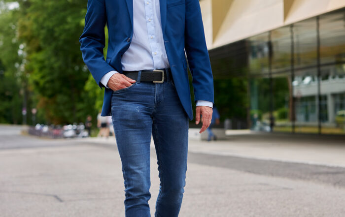 Ein Mann in blauem Blazer und Jeans schreitet selbstbewusst eine Stadtstraße entlang. Die moderne Kulisse mit großen Fenstern und von Bäumen gesäumten Gehwegen erinnert an den lebendigen Campus der Hochschule Coburg. Er lächelt und strahlt Entspannung und Gelassenheit aus.