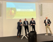 Vier Männer in Anzügen stehen auf einer Bühne in der Hochschule Coburg. Drei Männer unterschreiben Dokumente auf einem Tisch. Im Hintergrund ist eine Präsentationsfolie zu sehen, auf der neben Text auch das Bild einer Person in einer Blumenwiese zu sehen ist.