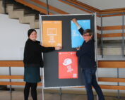 Zwei Personen hängen bunte Plakate an ein schwarzes Brett der Hochschule Coburg. Die mit Text und Bildern gefüllten Plakate stehen vor einer Treppe mit Holzgeländer in einem hellen Raum. Sie lächeln in die Kamera und fangen die lebendige Atmosphäre des Campus ein.