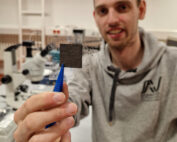 In einem Labor, in dem Spitzenforschung betrieben wird, manipuliert eine Person in einem grauen Kapuzenpullover vorsichtig ein quadratisches schwarzes Objekt mit einer Pinzette. Die makellose Umgebung voller Mikroskope und hochmoderner Geräte spiegelt den Innovationsgeist der Hochschule Coburg wider.