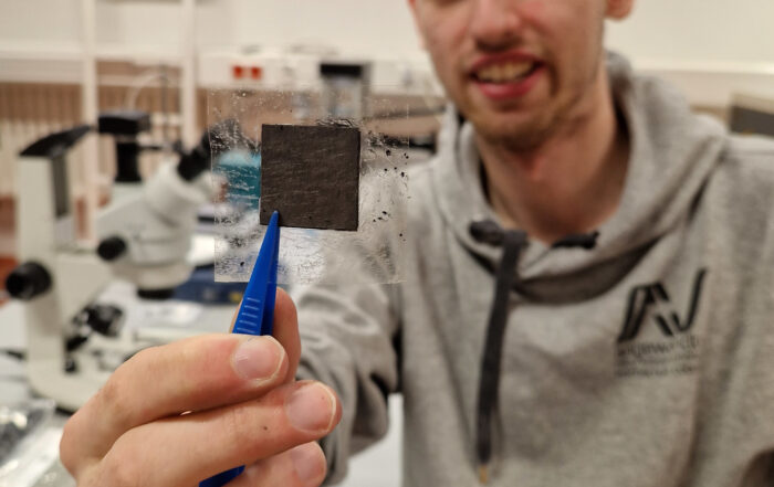 In einem Labor, in dem Spitzenforschung betrieben wird, manipuliert eine Person in einem grauen Kapuzenpullover vorsichtig ein quadratisches schwarzes Objekt mit einer Pinzette. Die makellose Umgebung voller Mikroskope und hochmoderner Geräte spiegelt den Innovationsgeist der Hochschule Coburg wider.