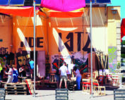 Eine lebhafte Szene im Freien entfaltet sich, wenn sich Menschen unter bunten Stoffdächern an der Hochschule Coburg versammeln. Holzstühle und Paletten sind auf dem Boden verteilt, während eine mit Graffiti verzierte Wand eine lebendige Kulisse bietet. Die Atmosphäre ist gesellig und entspannt und lädt alle ein, die lebhafte Umgebung zu genießen.