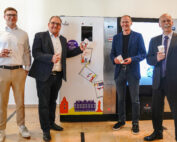 Vier Männer in Business-Kleidung posieren mit Bechern vor einem Getränkeautomaten an der Hochschule Coburg. Der geflieste Boden und die farbenfrohen Bechergrafiken auf dem Automaten verleihen dieser College-Szene einen modernen Touch.