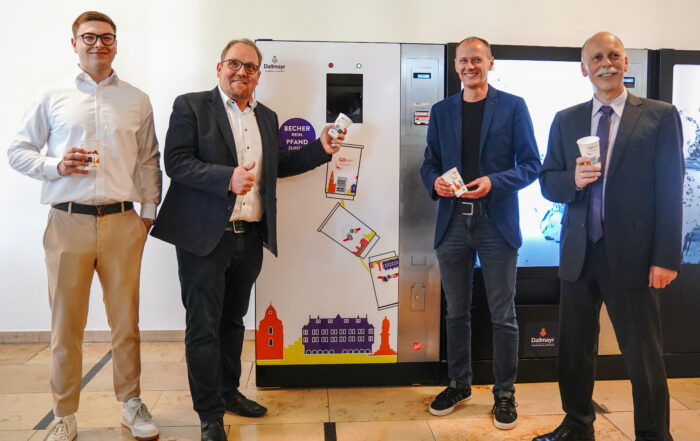 Vier Männer in Business-Kleidung posieren mit Bechern vor einem Getränkeautomaten an der Hochschule Coburg. Der geflieste Boden und die farbenfrohen Bechergrafiken auf dem Automaten verleihen dieser College-Szene einen modernen Touch.