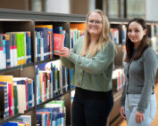 Zwei Frauen stehen vor den Bücherregalen der Bibliothek der Hochschule Coburg. Eine Frau mit Brille sucht sich ein Buch aus, die andere lächelt in die Kamera. Die Bibliothek ist gut beleuchtet, die Regale sind mit bunten Büchern gefüllt.