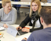 Drei Personen sitzen an einem Tisch in einem Hörsaal der Hochschule Coburg und diskutieren. Eine Person benutzt einen Laptop, auf dem Tisch liegen Papiere und Notizbücher. Der Raum hat weiße Wände und im Hintergrund Schreibtische.