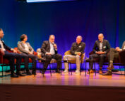 Sechs Personen sitzen auf einer Bühne der Hochschule Coburg und nehmen an einer Podiumsdiskussion teil. Sie tragen Business-Kleidung und halten Mikrofone in der Hand und tragen so zum formellen Rahmen bei. Der Hintergrund ist blau und sanft beleuchtet, was eine professionelle Atmosphäre schafft.