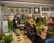 Eine Gruppe von Menschen, vermutlich von der Hochschule Coburg, lächelt und hält Pflanzen vor einem Schaufenster mit dem Schild „Raum für Zukunftsideen“. Die Szene zeigt eine Vielzahl von Topfkräutern und Blumen auf einem Holztisch.