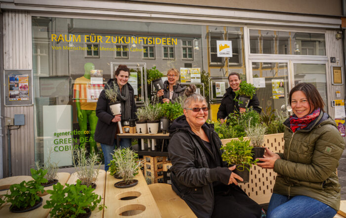 Eine Gruppe von Menschen, vermutlich von der Hochschule Coburg, lächelt und hält Pflanzen vor einem Schaufenster mit dem Schild „Raum für Zukunftsideen“. Die Szene zeigt eine Vielzahl von Topfkräutern und Blumen auf einem Holztisch.