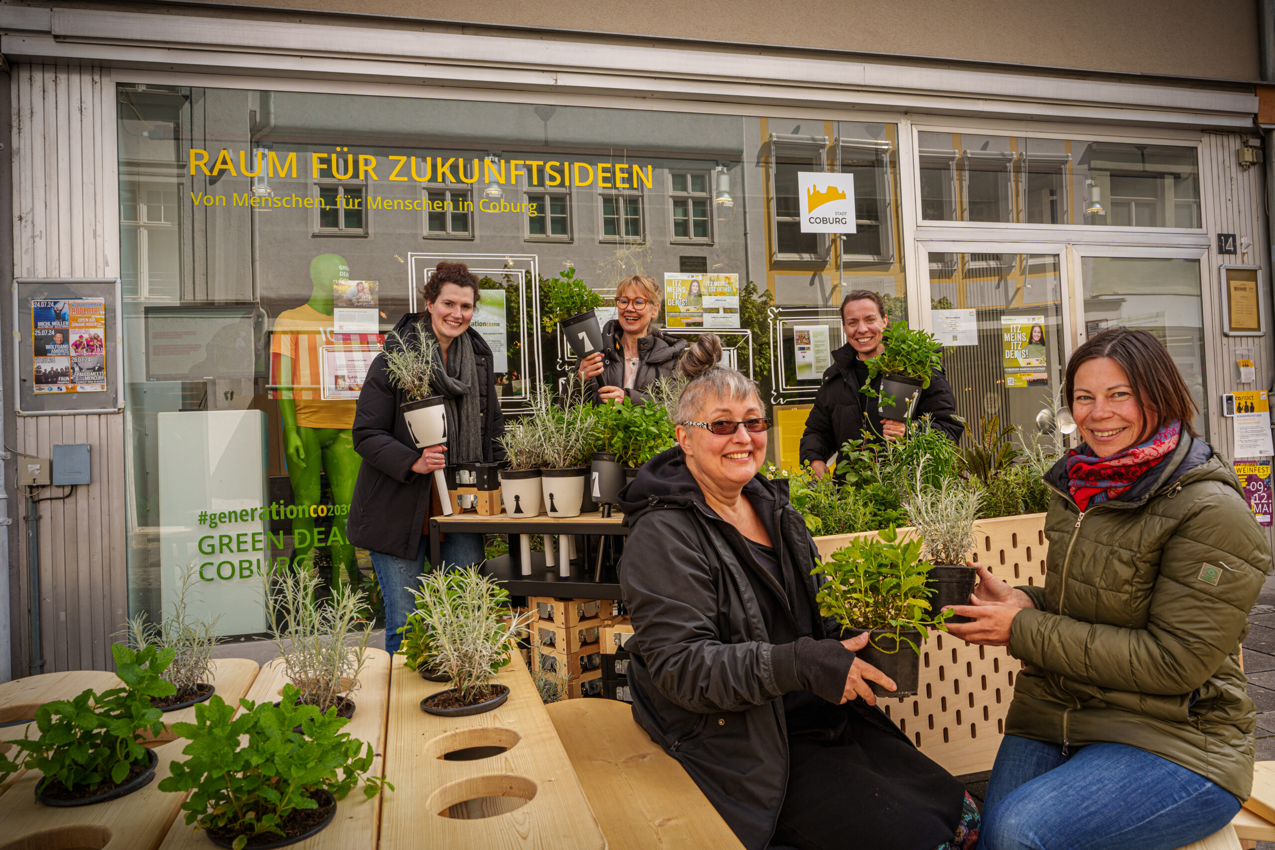 Eine Gruppe von Menschen, vermutlich von der Hochschule Coburg, lächelt und hält Pflanzen vor einem Schaufenster mit dem Schild „Raum für Zukunftsideen“. Die Szene zeigt eine Vielzahl von Topfkräutern und Blumen auf einem Holztisch.