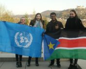 Vier Menschen lächeln und halten Flaggen im Freien: Einer hält eine Flagge der Vereinten Nationen, während andere stolz die Flagge des Südsudan zeigen. Vor der malerischen Kulisse von Hügeln und Gebäuden feiern diese Studenten der Hochschule Coburg Einheit und globale Bildung.