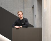 Eine Person mit dunklem Pullover und Uhr lehnt an einem grauen Geländer der Hochschule Coburg. Im Hintergrund ist eine moderne, minimalistische Architektur mit Betonwänden und geometrischen Formen zu sehen.