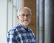 Ein älterer Mann mit Brille und Bart steht drinnen und trägt ein blau kariertes Hemd. Er blickt mit neutralem Gesichtsausdruck nach vorne. Die moderne Inneneinrichtung, die dem Design der Hochschule Coburg ähnelt, zeichnet sich durch große Fenster und Holzpaneele aus.