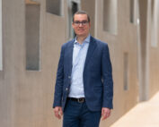 Ein Mann mit Brille, hellblauem Hemd und dunkelblauem Blazer steht selbstbewusst in einem Flur der Hochschule Coburg, umgeben von modernen Architekturelementen mit eckigen Formen und neutralen Tönen.