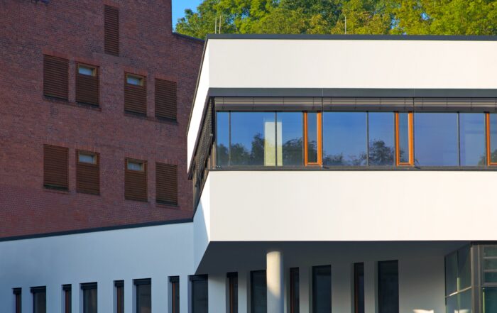 Das moderne Gebäude der Hochschule Coburg weist einen markanten Kontrast auf: ein weißer, eckiger Baukörper mit großen Fenstern vor einer älteren, roten Backsteinfassade. Im Hintergrund gedeihen üppig grüne Bäume unter einem klaren blauen Himmel.