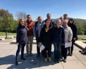 Eine Gruppe von neun Personen, möglicherweise Studenten der Hochschule Coburg, steht an einem sonnigen Tag im Freien zusammen und lächelt für das Foto. Sie sind in legerer Kleidung gekleidet und von Bäumen und einem klaren blauen Himmel umgeben.