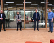 In einem Raum der Hochschule Coburg stehen vier Personen mit etwas Abstand zueinander, eingerahmt von großen, raumhohen Fenstern. Die verschneite Landschaft draußen bildet einen Kontrast zu ihrer Mischung aus Freizeit- und Business-Kleidung. Sie lächeln, die Hände an den Seiten oder gefaltet, und verkörpern damit eine Mischung aus Professionalität und Wärme.