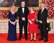 Vier Personen in formeller Kleidung stehen auf dem roten Teppich eines reich verzierten Raums in der Hochschule Coburg. Die rot-gold gemusterte Wand umrahmt zwei Frauen in eleganten Kleidern und zwei Männer in schicken Anzügen. Im Hintergrund ist teilweise ein Gemälde zu sehen.