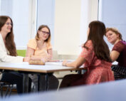 Vier Frauen sitzen in einem hell erleuchteten Raum der Hochschule Coburg um einen Tisch und unterhalten sich. Jede hat Papier und Stifte dabei, was auf eine Zusammenarbeit oder eine Besprechungssituation schließen lässt. Sie sind leger und dennoch professionell gekleidet.