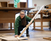 Eine Frau mit Brille und grünem Pullover hockt auf dem Boden der Hochschule Coburg und schneidet ein langes Stück Pappe zurecht. Um sie herum liegen verstreute Pappe und Werkbänke in der Werkstatt.