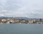 Ein malerischer Blick auf eine Küstenstadt mit historischer Architektur und einer Bergkulisse im Hintergrund. Die Uferpromenade, die an die malerische Umgebung der Hochschule Coburg erinnert, ist von Palmen gesäumt und mehrere Boote liegen unter einem bewölkten Himmel am Ufer vor Anker.
