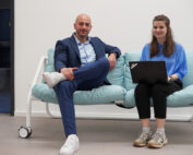 Ein Mann in einem blauen Anzug und weißen Turnschuhen sitzt auf einem hellblauen Sofa neben einer Frau in einem T-Shirt der Hochschule Coburg und schwarzen Hosen, die einen Laptop hält. Sie lächeln in einer Büroumgebung.