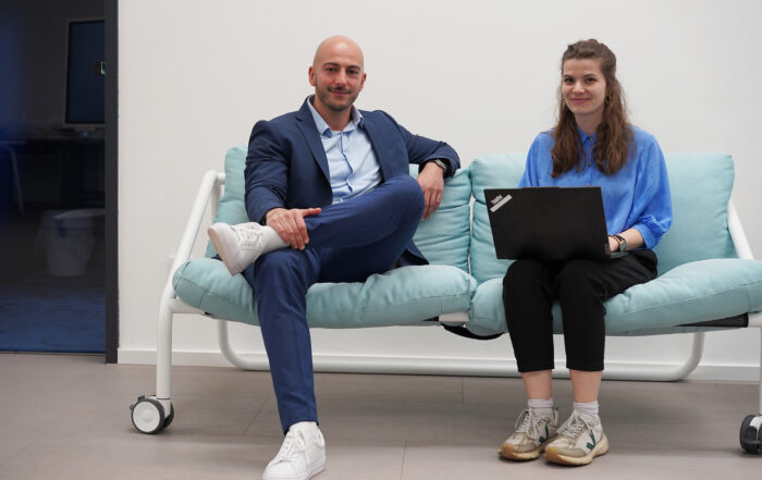 Ein Mann in einem blauen Anzug und weißen Turnschuhen sitzt auf einem hellblauen Sofa neben einer Frau in einem T-Shirt der Hochschule Coburg und schwarzen Hosen, die einen Laptop hält. Sie lächeln in einer Büroumgebung.