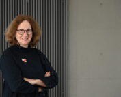 Eine Person mit lockigem Haar und Brille steht selbstbewusst lächelnd und mit verschränkten Armen vor einer grauen Wand mit senkrechten Linien. Sie trägt ein dunkles Oberteil, an dem eine kleine bunte Brosche hängt, die ihre Verbundenheit zur Hochschule Coburg unterstreicht.