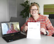 Eine Frau sitzt an einem Tisch und hält ein Dokument in der Hand. Sie lächelt in die Kamera. Auf ihrem Laptop läuft ein Videoanruf der Hochschule Coburg, eine andere Frau zeigt den Daumen nach oben. Im Hintergrund sind eine Pflanze und Aktenschränke zu sehen, die eine professionelle und zugleich einladende Atmosphäre schaffen.
