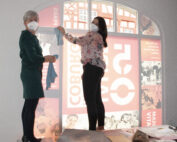 Zwei maskierte Personen stehen auf einem Tisch und hängen ein Tuch mit grafischen Motiven an ein großes Innenfenster der Hochschule Coburg. Die eine trägt ein grünes Kleid, die andere ein geblümtes Hemd und verleiht dem Arbeits- oder Ausstellungsbereich ein künstlerisches Flair.