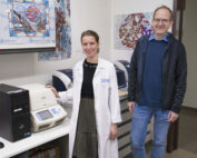 Eine Frau im Laborkittel steht neben einem Mann in Freizeitkleidung, beide lächelnd, an der Hochschule Coburg. Sie sind umgeben von Geräten und bunten wissenschaftlichen Postern an den Wänden, die ihre lebendige Forschungsumgebung widerspiegeln.