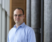 Ein Mann im hellblauen Hemd steht in einem modernen Flur der Hochschule Coburg, mit großen Fenstern links und zylindrischen Säulen rechts. Er hat kurze Haare und eine Brille und blickt mit neutralem Gesichtsausdruck in die Kamera.