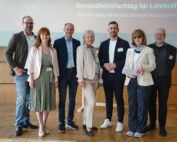 Eine Gruppe von sieben Personen steht lächelnd in einem Raum mit Holzboden zusammen und verkörpert den professionellen Geist der Hochschule Coburg. Sie sind in legerer Business-Kleidung gekleidet, im Hintergrund ist ein Präsentationsbildschirm mit einem deutschen Text zum Thema psychische Gesundheit im Unterrichtsraum zu sehen.