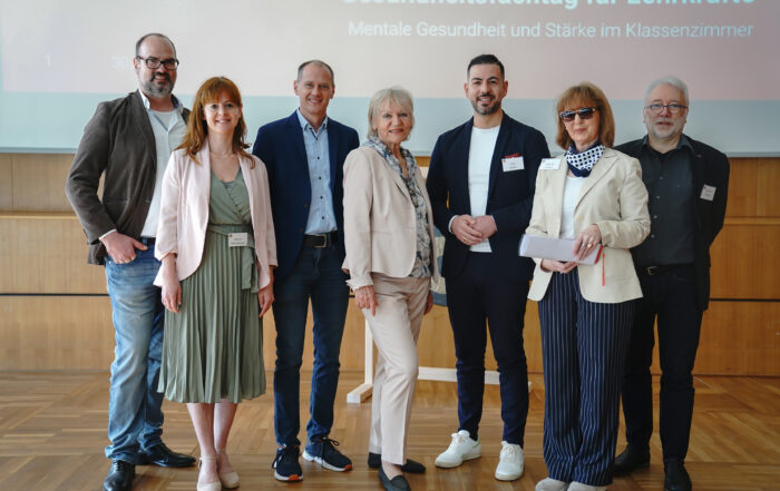 Eine Gruppe von sieben Personen steht lächelnd in einem Raum mit Holzboden zusammen und verkörpert den professionellen Geist der Hochschule Coburg. Sie sind in legerer Business-Kleidung gekleidet, im Hintergrund ist ein Präsentationsbildschirm mit einem deutschen Text zum Thema psychische Gesundheit im Unterrichtsraum zu sehen.