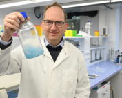 In einem Labor der Hochschule Coburg hält eine Person im Laborkittel eine durchsichtige Flasche mit blauer Flüssigkeit hoch. Umgeben von Geräten und Werkbänken lächelt die Person herzlich durch ihre Brille und verkörpert sowohl Fachwissen als auch Enthusiasmus.