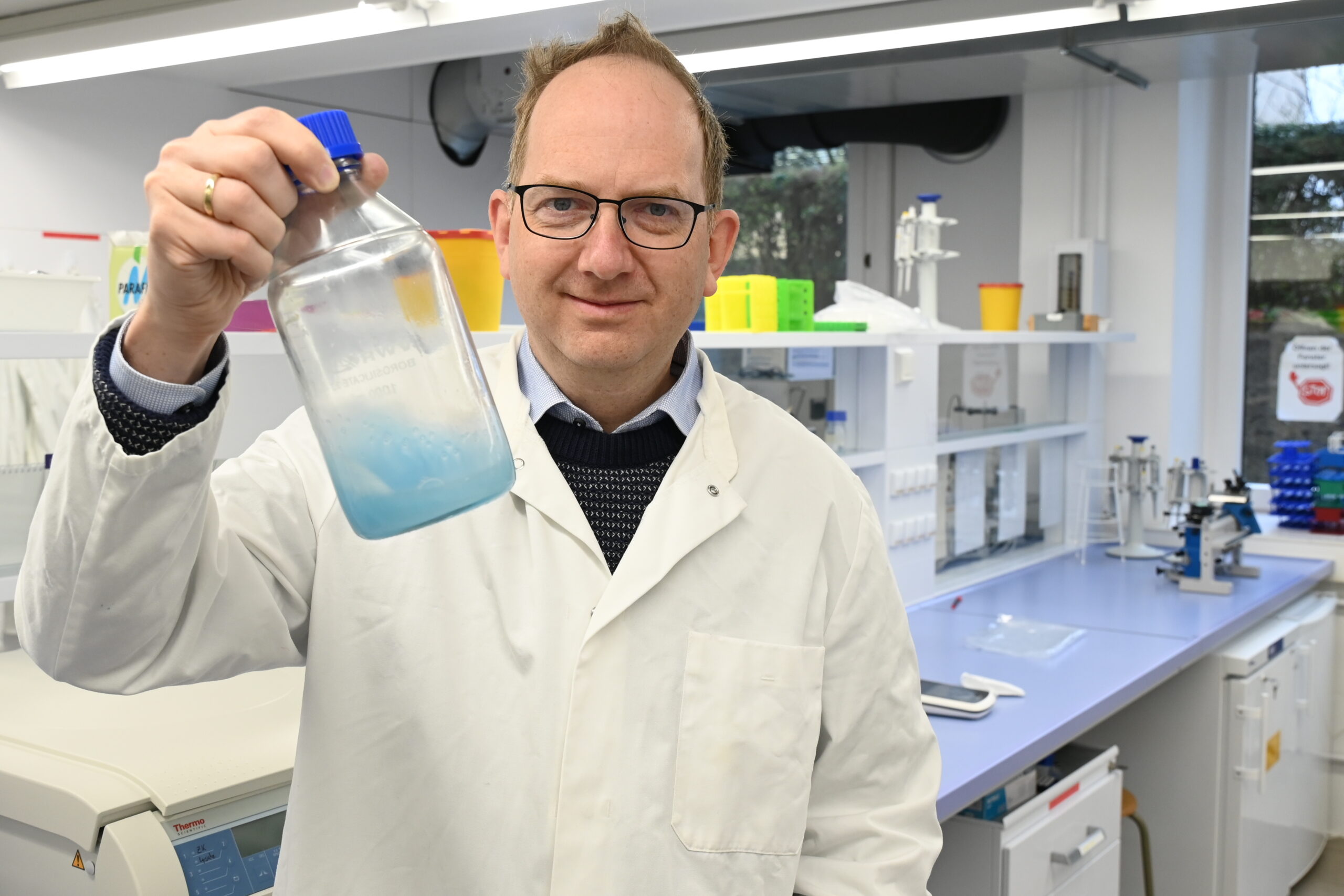 In einem Labor der Hochschule Coburg hält eine Person im Laborkittel eine durchsichtige Flasche mit blauer Flüssigkeit hoch. Umgeben von Geräten und Werkbänken lächelt die Person herzlich durch ihre Brille und verkörpert sowohl Fachwissen als auch Enthusiasmus.