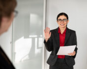 Eine Frau im Anzug hält ein Dokument und hebt die Hand. Es sieht so aus, als würde sie einen Eid ablegen oder eine Präsentation an der Hochschule Coburg halten. Sie steht in einem hell erleuchteten Büro mit Milchglasscheiben im Hintergrund. Im Vordergrund ist eine weitere Person unscharf zu sehen, ihr gegenüber.