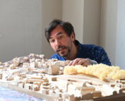 An der Hochschule Coburg beugt sich ein Mann über ein detailliertes Architekturmodell einer Stadtlandschaft. Das Modell zeigt kleine Gebäude und Strukturen aus Holz, deren Abschnitte an Bäume oder Grünpflanzen erinnern und aus strukturiertem gelbem Material gefertigt sind.