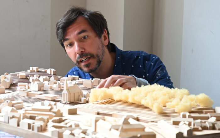 An der Hochschule Coburg beugt sich ein Mann über ein detailliertes Architekturmodell einer Stadtlandschaft. Das Modell zeigt kleine Gebäude und Strukturen aus Holz, deren Abschnitte an Bäume oder Grünpflanzen erinnern und aus strukturiertem gelbem Material gefertigt sind.
